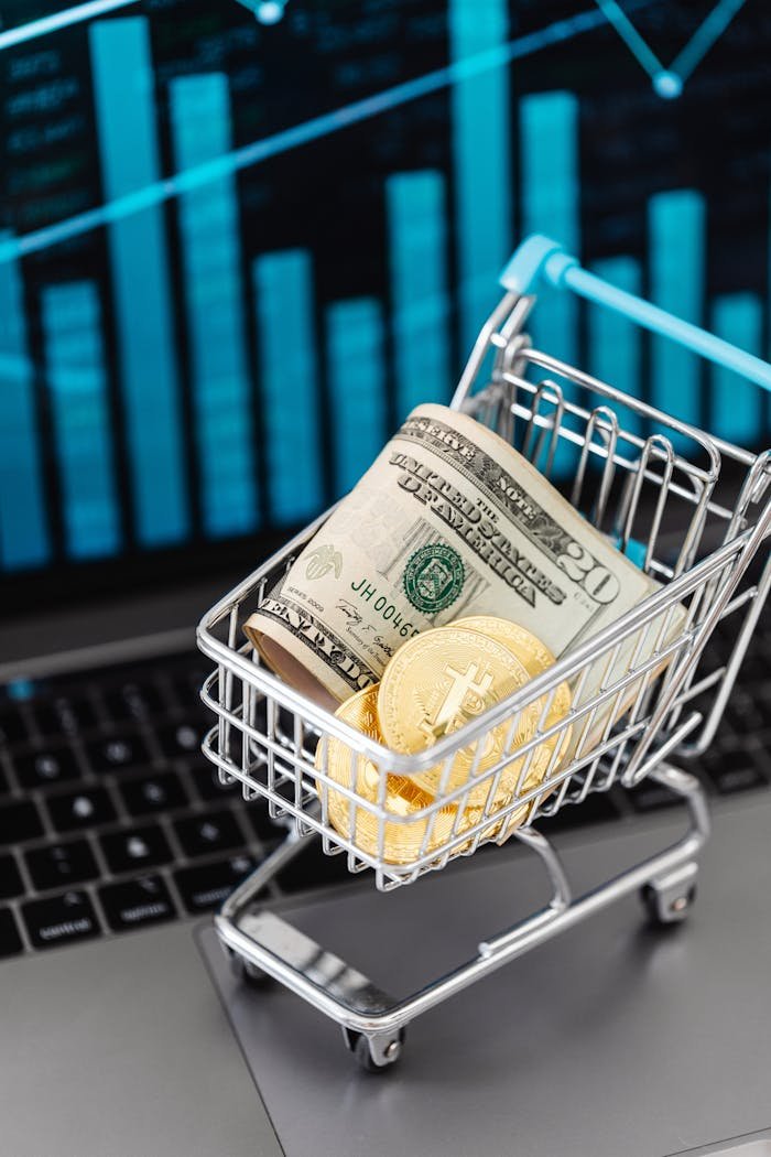 Shopping cart with bitcoin and cash on laptop with financial graph backdrop.